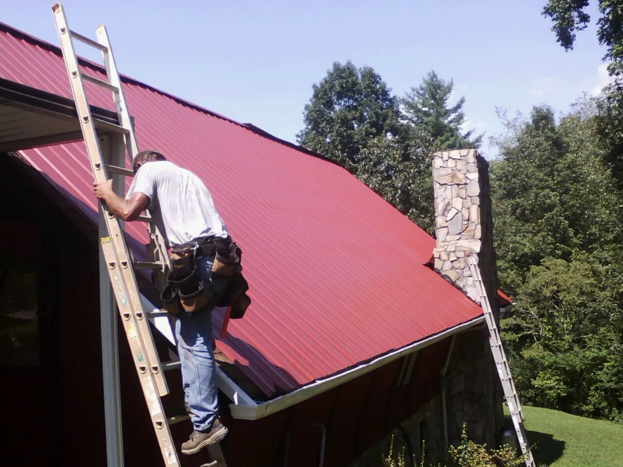 Roofer on a ladder beside a red metal roof and stone chimney — Soffit & Fascia Repair by Roofing Brothers in Stuart